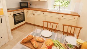 Une cuisine avec des meubles blancs, un four à micro-ondes, un four traditionnel, une fenêtre donnant sur un paysage verdoyant, et une table à manger dressée avec du pain, du fromage et des légumes.