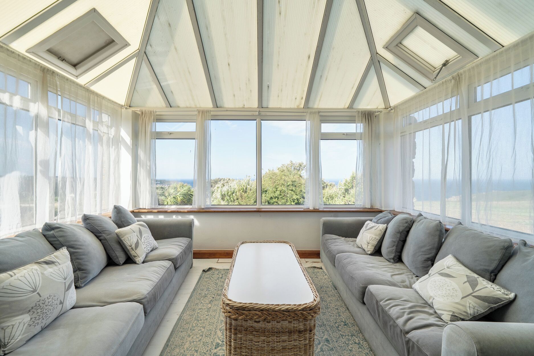 A sunroom with two grey sofas, a central table, and large windows offering a view of greenery.
