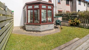 Una solana en esquina con base de piedra y paneles de vidrio, contigua a una casa blanca con detalles en marrón y una valla de madera.