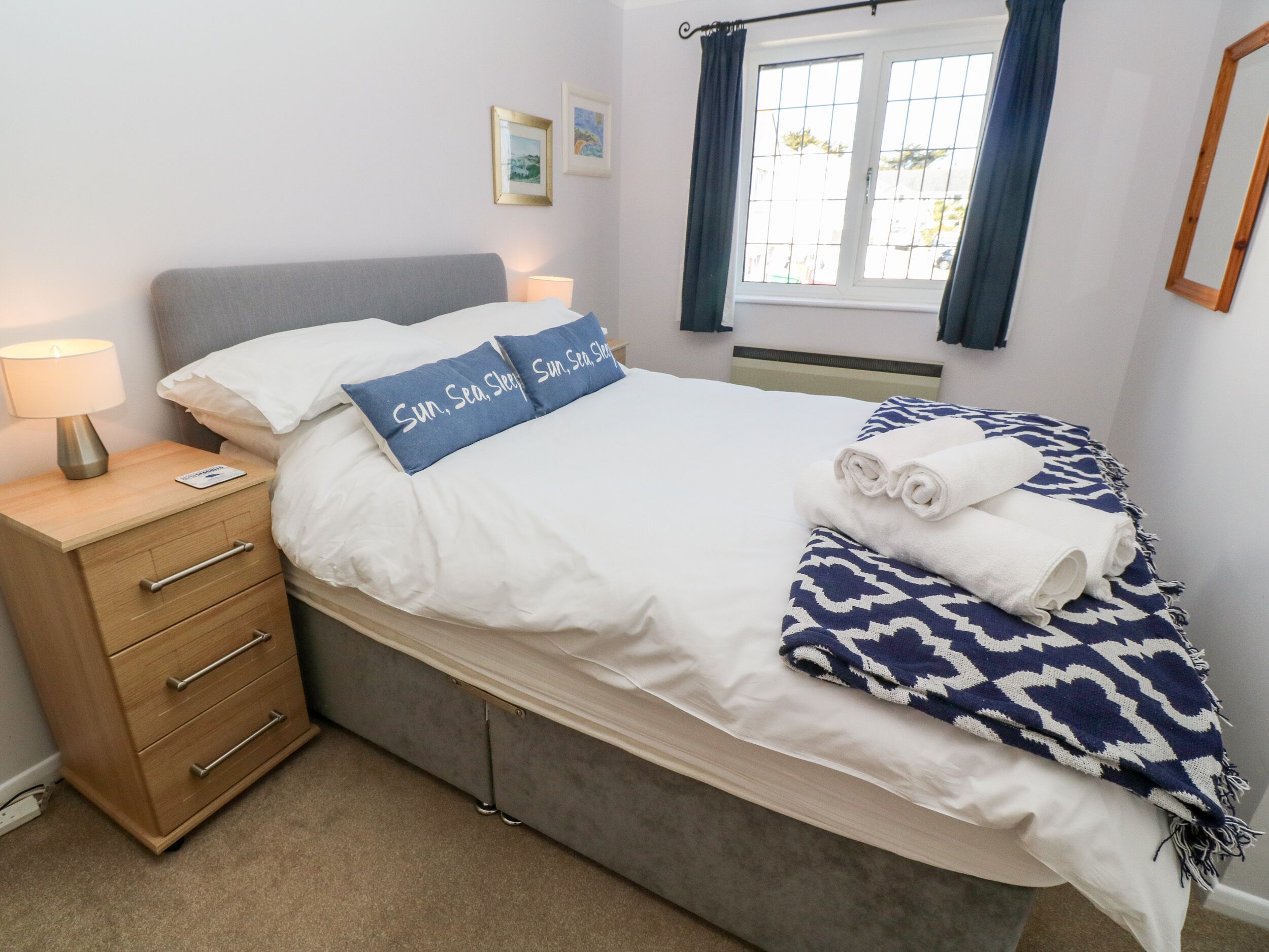 A neatly made bed with white bedding and a blue pillow with text, a bedside lamp, a wooden nightstand with drawers, and a window with curtains.