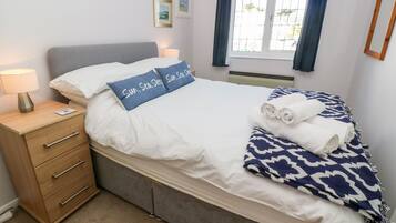A neatly made bed with white bedding and a blue pillow with text, a bedside lamp, a wooden nightstand with drawers, and a window with curtains.