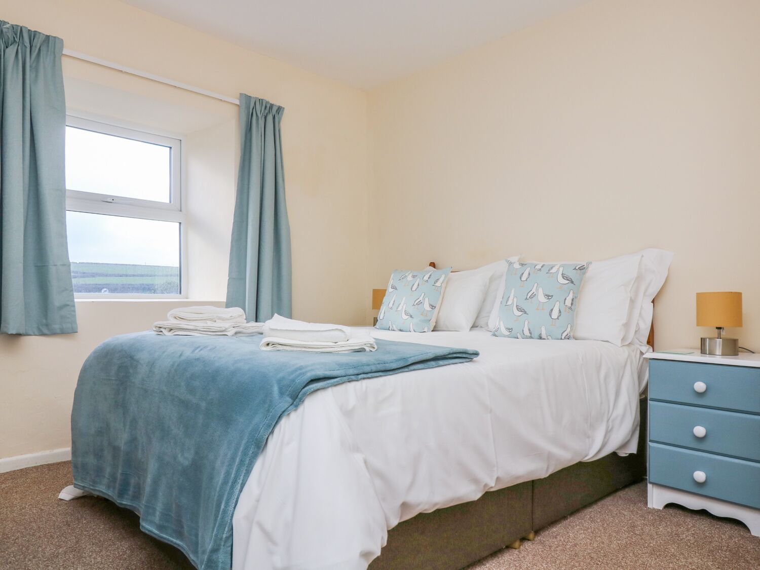 A neatly made bed with a blue blanket, white pillows, and a matching blue headboard. There is a window with curtains, a bedside lamp, and a small drawer unit beside the bed.