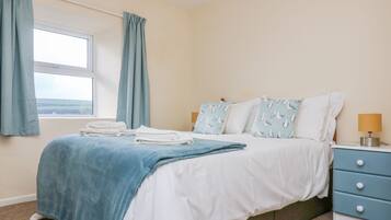 A neatly made bed with a blue blanket, white pillows, and a matching blue headboard. There is a window with curtains, a bedside lamp, and a small drawer unit beside the bed.