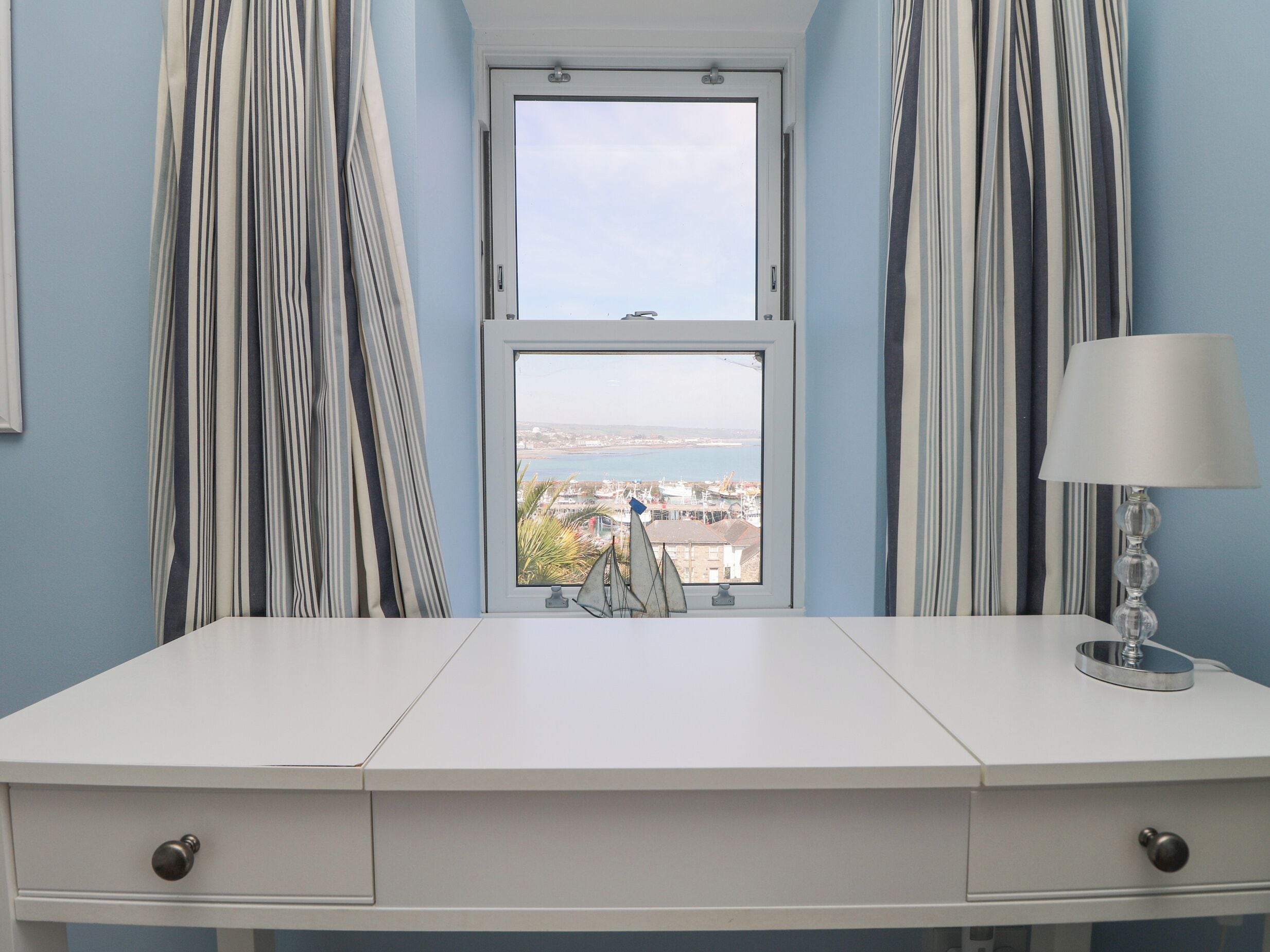 Un escritorio blanco con dos cajones, una ventana que ofrece vistas a una ciudad costera y cortinas a rayas.