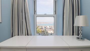 Un escritorio blanco con dos cajones, una ventana que ofrece vistas a una ciudad costera y cortinas a rayas.