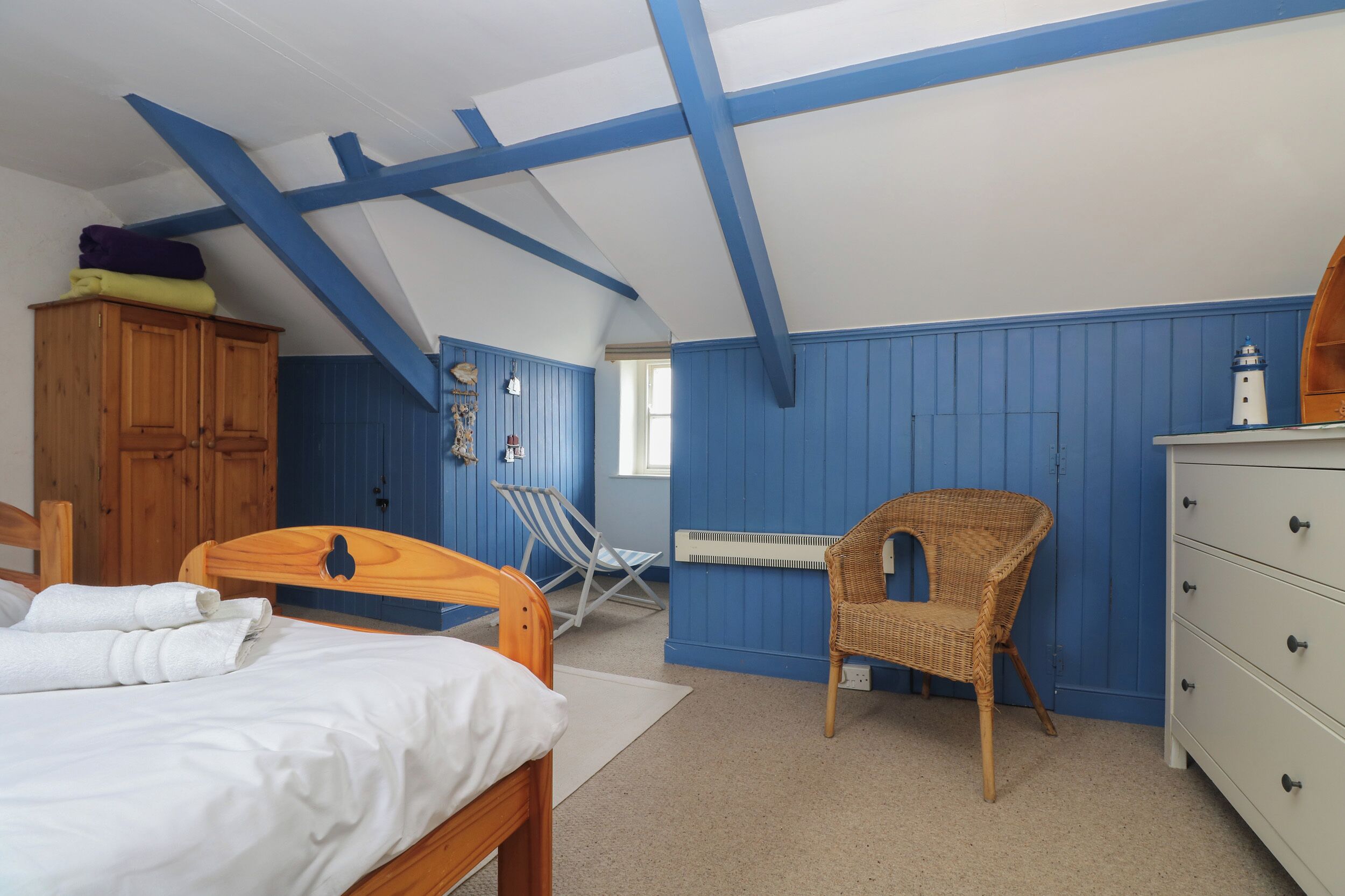 Una stanza con un letto, un'armadietto in legno, una sedia in vimini, un comò bianco e un termosifone.