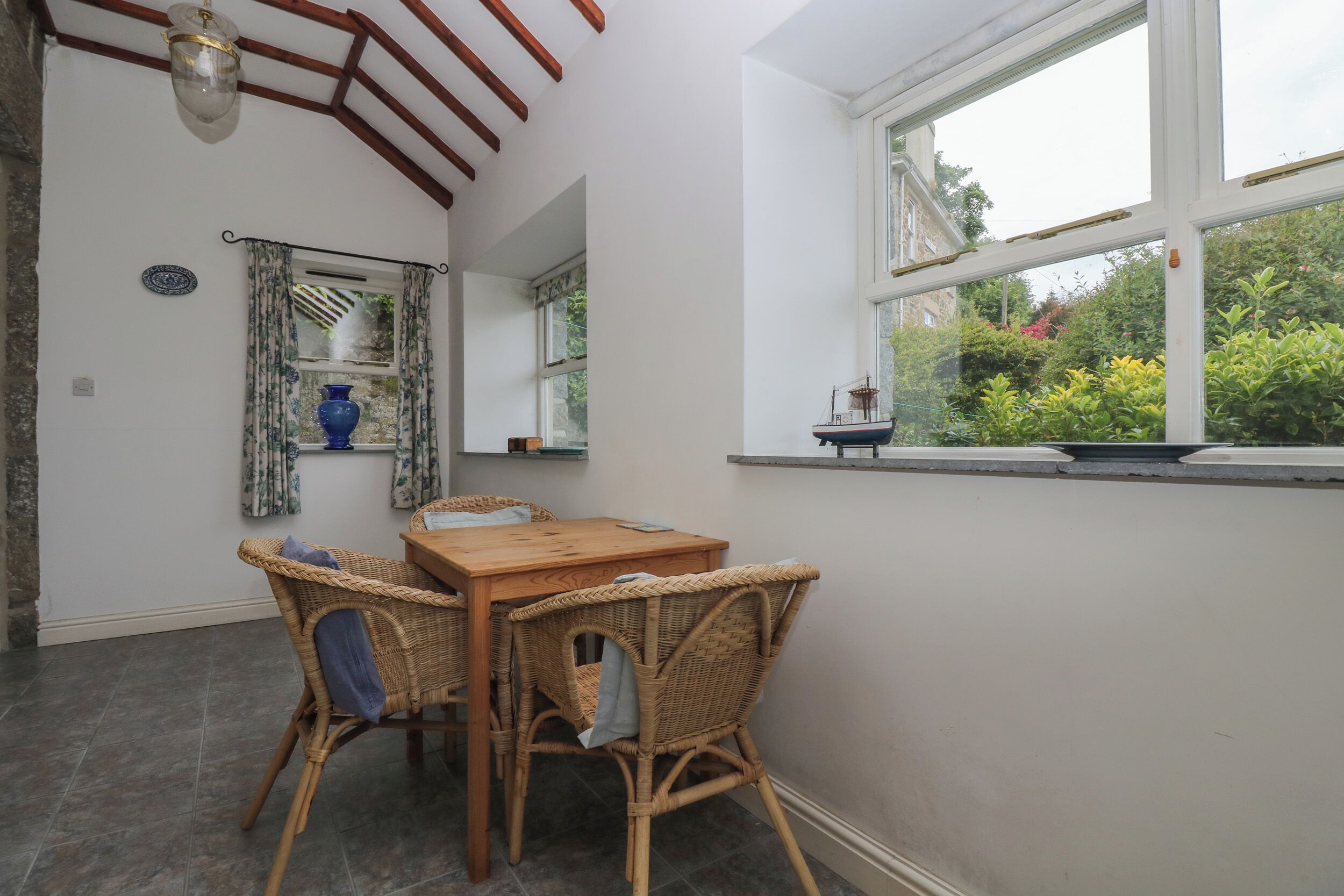 Una habitación con una mesa de madera y sillas de mimbre, una ventana con vistas a la vegetación y un jarrón decorativo en el alféizar.