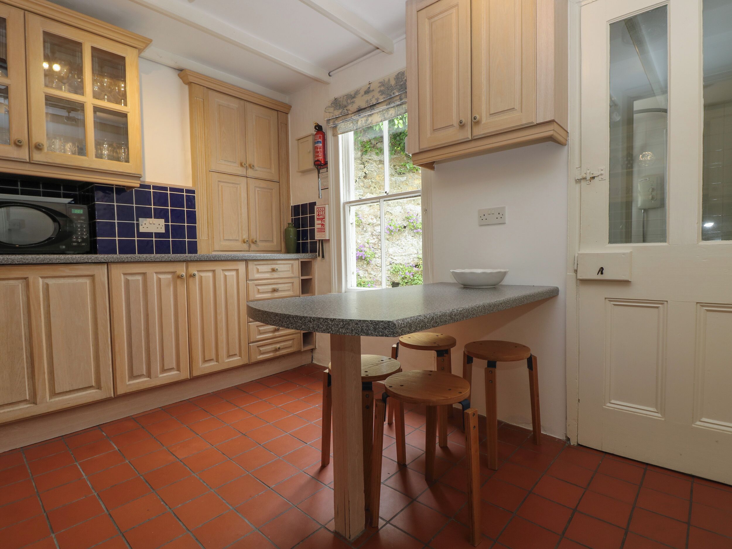 Een keuken met houten kasten, een magnetron, een spoelbak en een raam met uitzicht op groen.