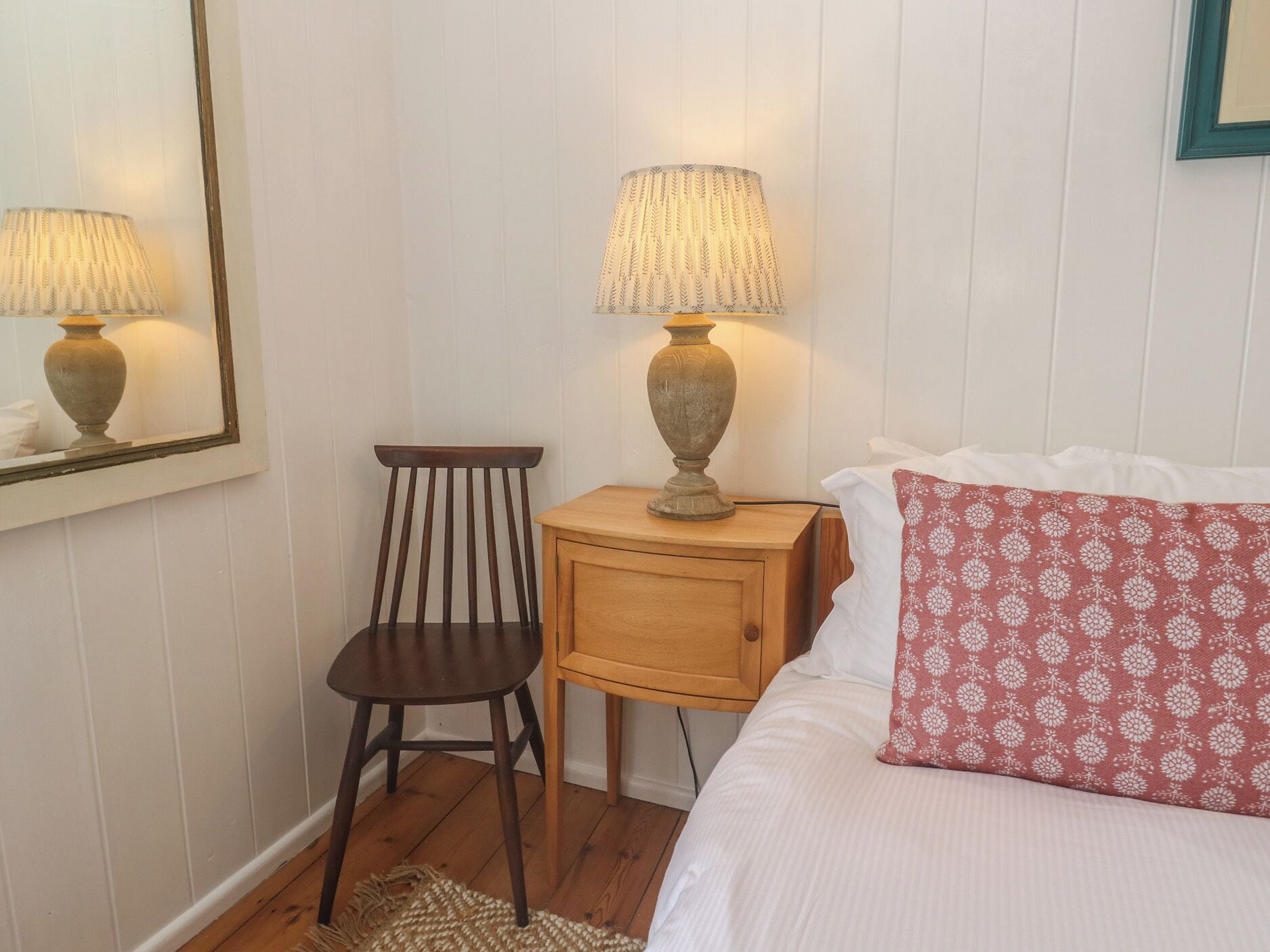 A bedroom with a wooden chair, a bedside table with a lamp, a bed with a patterned pillow, and a mirror on the wall.