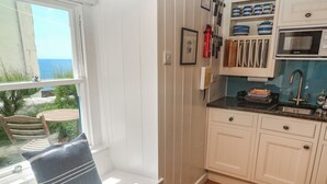 A kitchen with white cabinets, a built-in oven, and a window overlooking a beach. - Pegs (Helston)