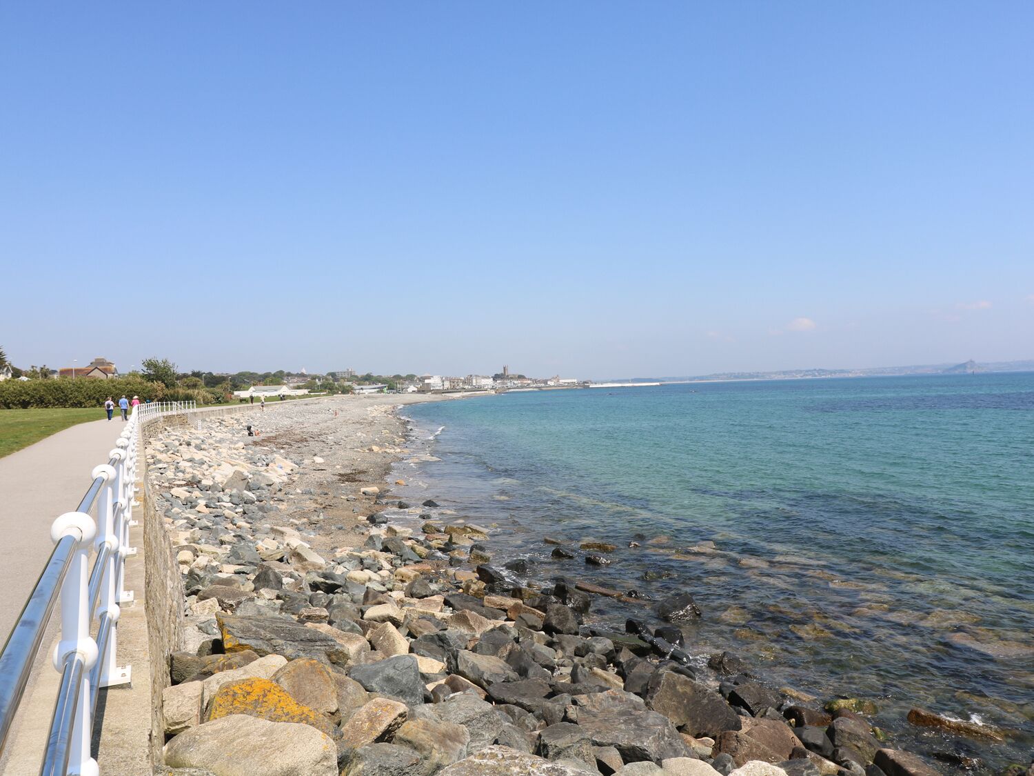 A rocky shoreline with a clear blue sea, a paved walkway, and buildings in the distance.