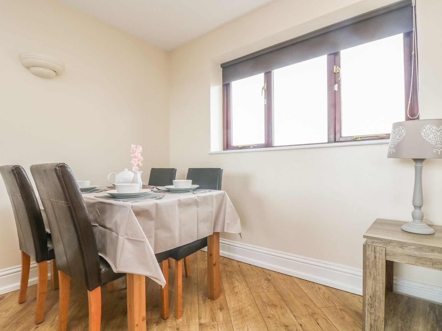 Comedor con una mesa preparada para cuatro, sillas de madera, una ventana con persianas y una lámpara sobre una mesita.