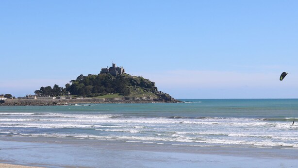 A coastal view with a castle on a hill, waves in the foreground, and a kite in the sky. - Seashells (Penzance)
