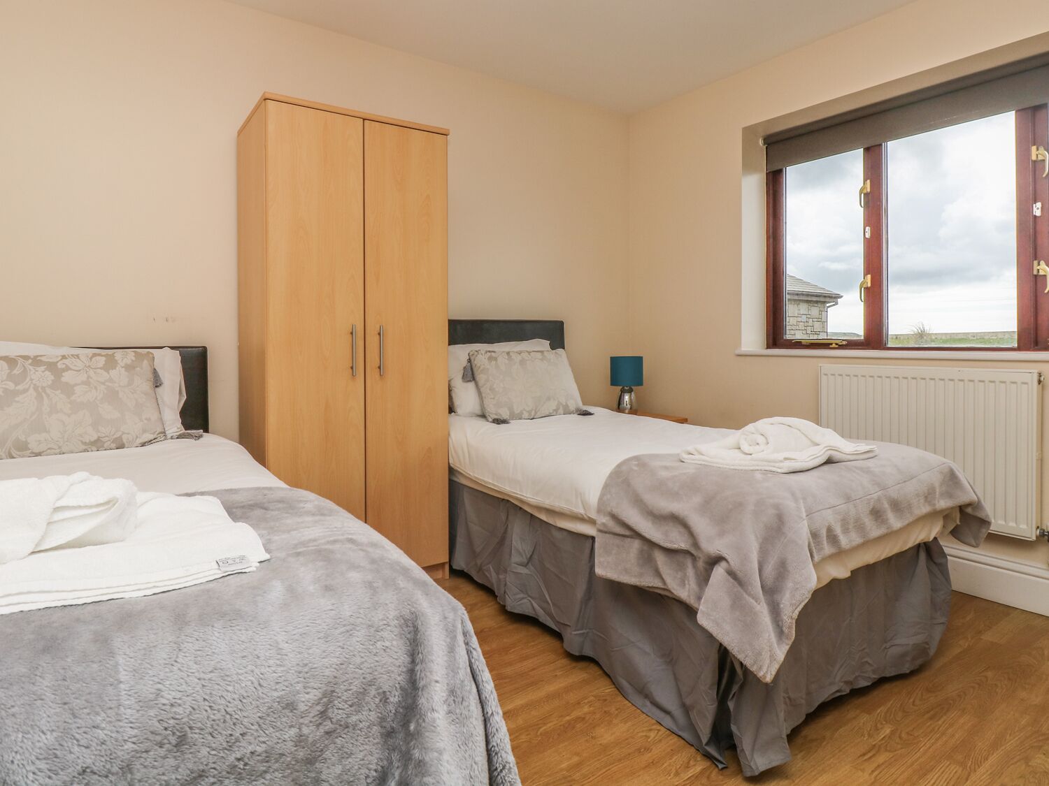 A bedroom with two beds, a wardrobe, a window with curtains, and a lamp.