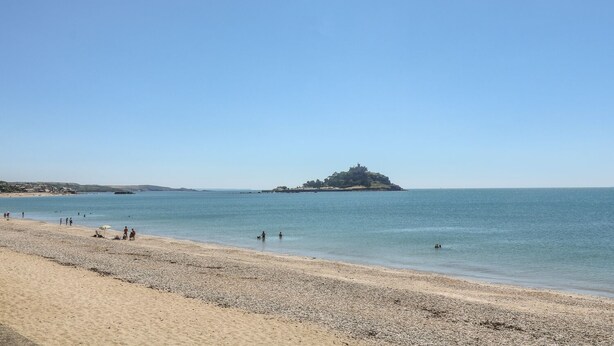 A beach with pebbles, a clear sky, and a distant island with buildings. - Seabreeze (Penzance)