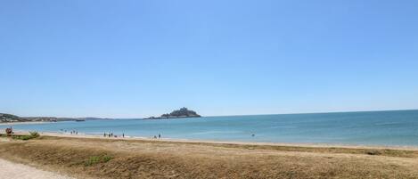 Una playa con un sendero de tierra que lleva al agua, una pequeña isla a lo lejos y un cielo azul despejado.