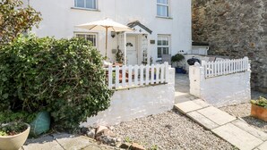 Une maison blanche à deux étages, avec une clôture en bois peinte en blanc, un petit jardin et un mur en pierre.