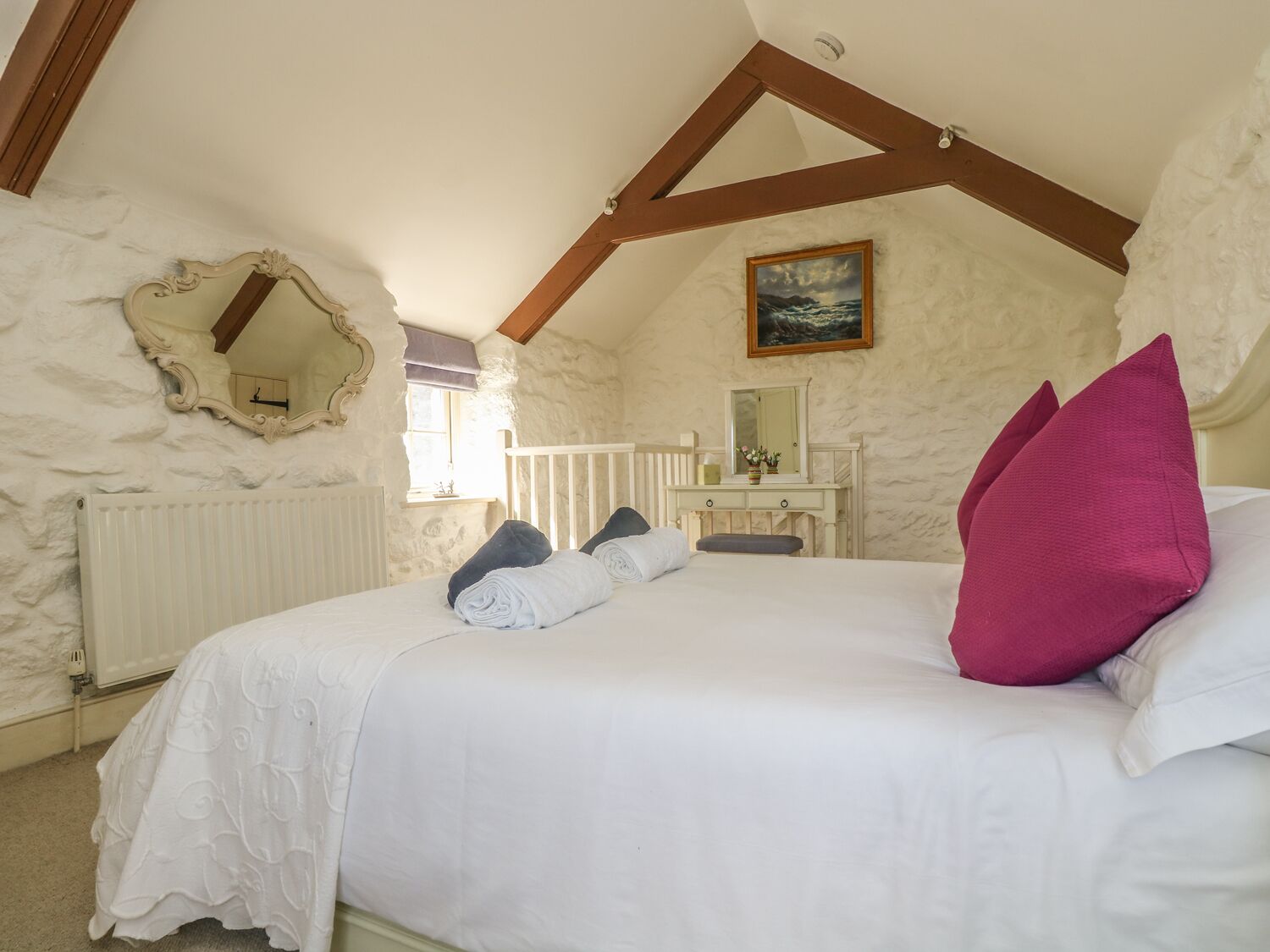 A bedroom with a sloped ceiling, a bed with white bedding and a pink pillow, a dresser with a mirror, and a painting on the wall.