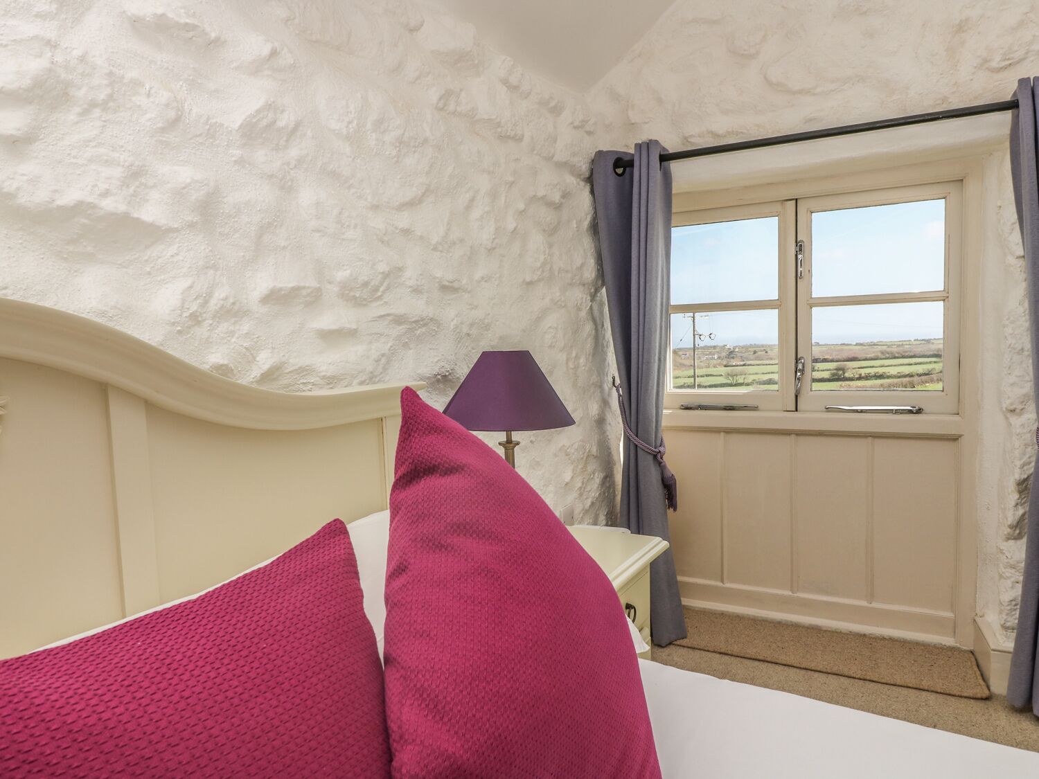 Een slaapkamer met een witte, gestructureerde muur, een bed met twee rode kussens, een nachtkastje met een lamp en een raam met uitzicht op het landschap.