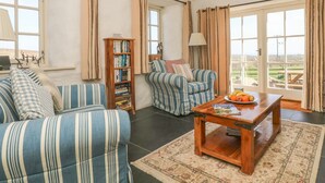 A living room with a sofa, armchair, and a wooden coffee table with fruit. There is a bookshelf and a view of the outdoors through large windows. - Boar's House (Penzance)