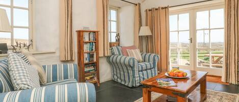 Una sala de estar con sofá, sillón y una mesa de centro de madera con fruta. Hay una estantería y vistas al exterior a través de amplias ventanas.
