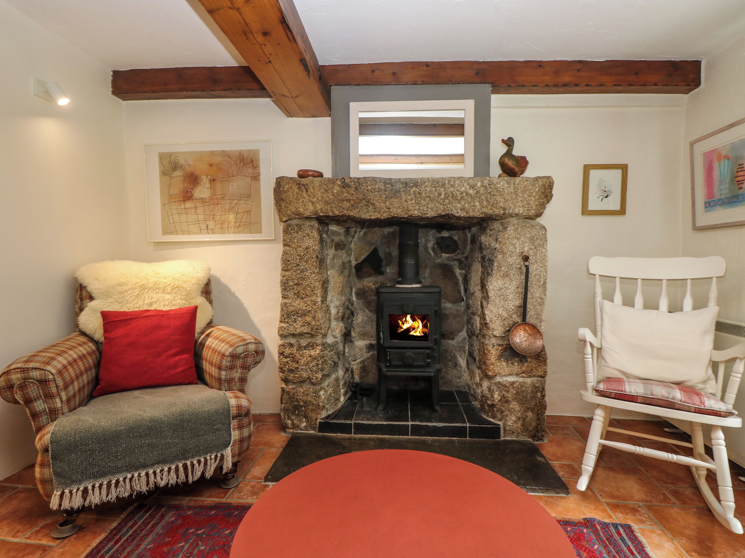 A cozy living room with a stone fireplace, a plaid armchair, a white rocking chair, and a red ottoman.