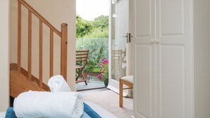 Une chambre avec un escalier en bois, un lit recouvert d’une couette à rayures bleues et blanches, une serviette blanche, et une vue sur un jardin par une porte ouverte.