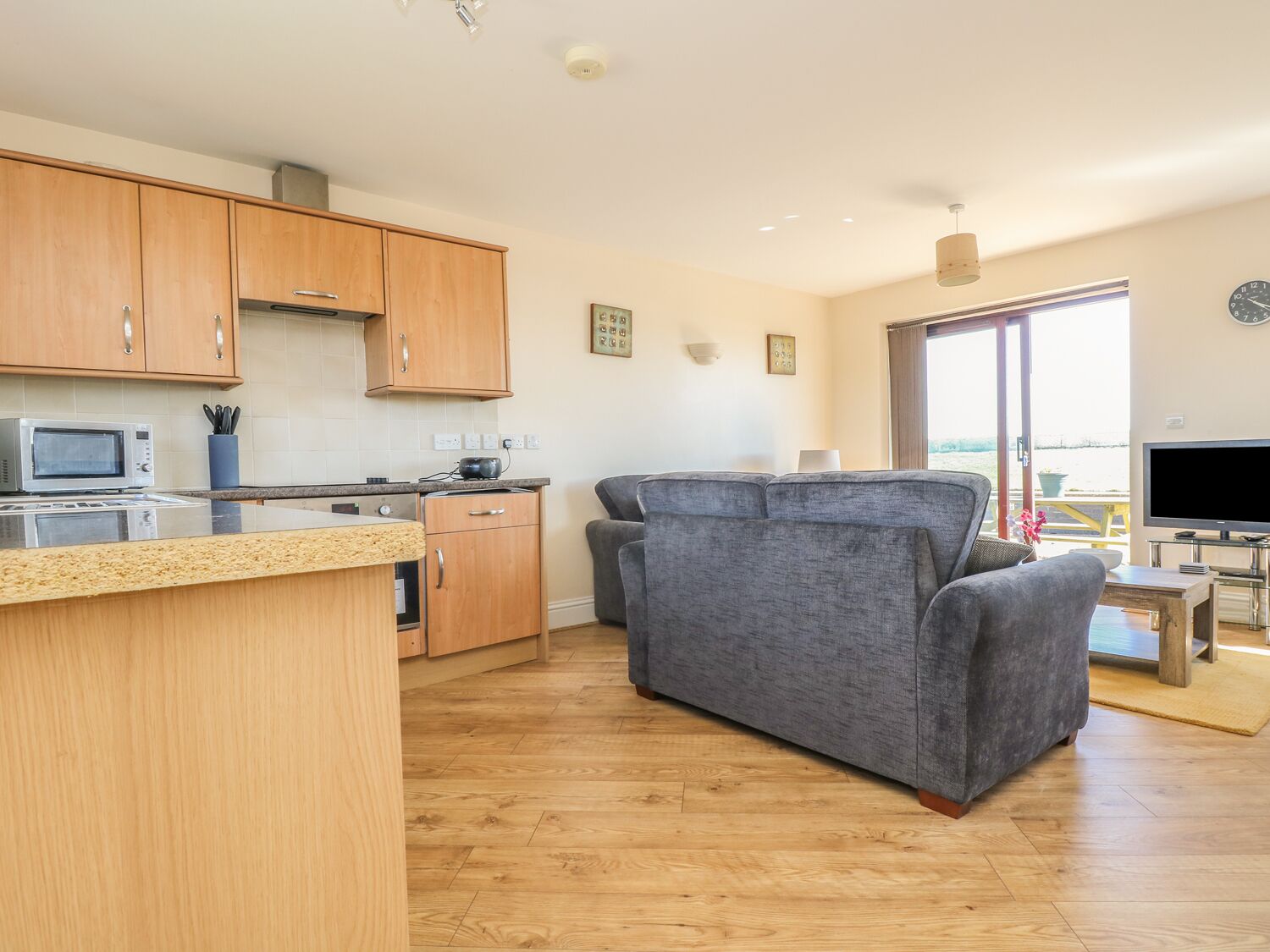 A modern kitchen with wooden cabinets, a microwave, and a small island. There is a grey sofa and a flat-screen TV in a living area with a large window.
