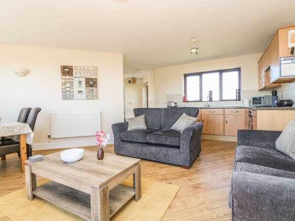 A modern living room with a grey sofa, a wooden coffee table, and a dining area with striped chairs. A kitchen with wooden cabinets and a window is visible in the background. - Surfside (Penzance)