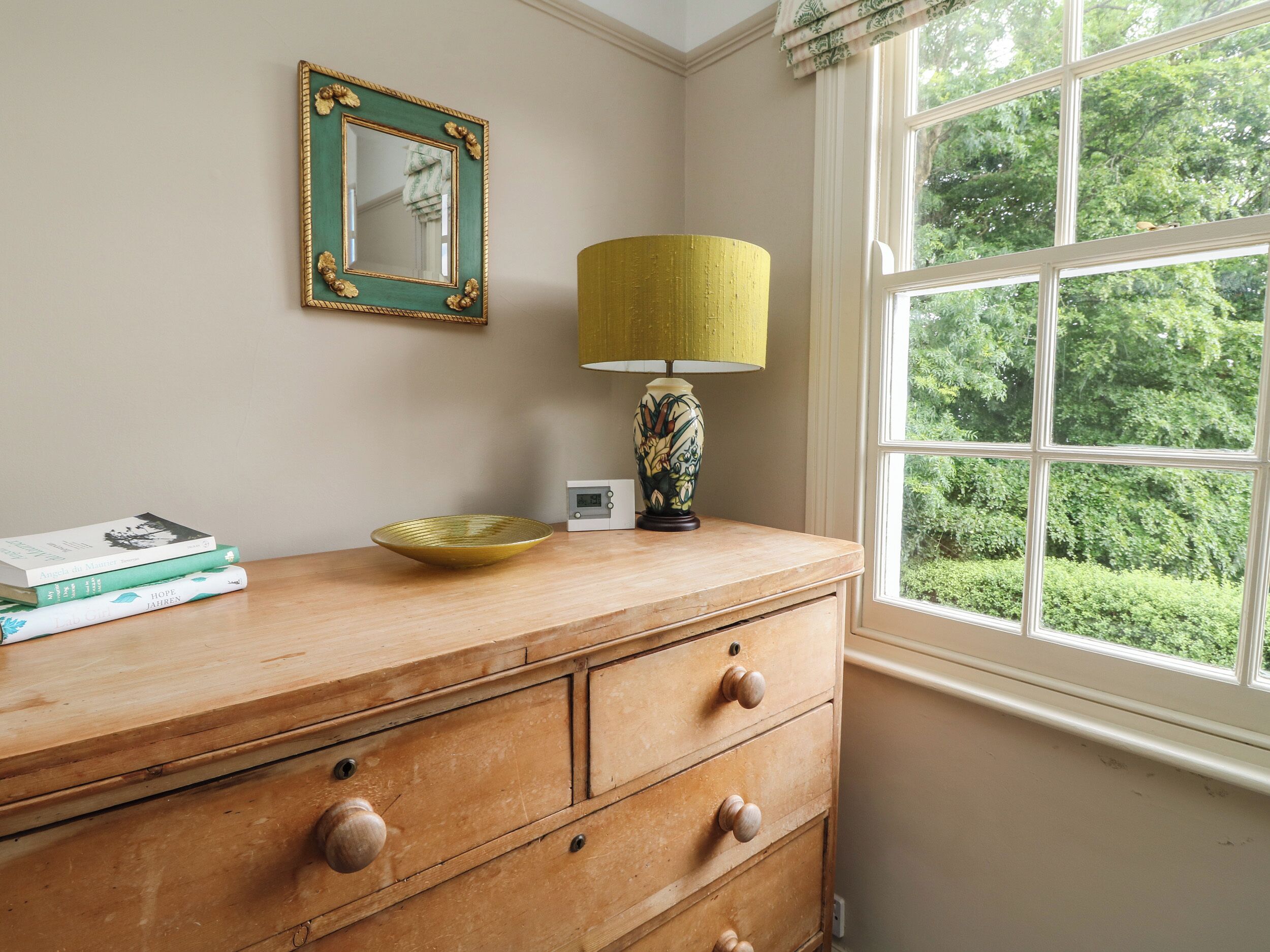 A wooden dresser with drawers, a green-framed mirror, a yellow lamp, and a bowl on top.