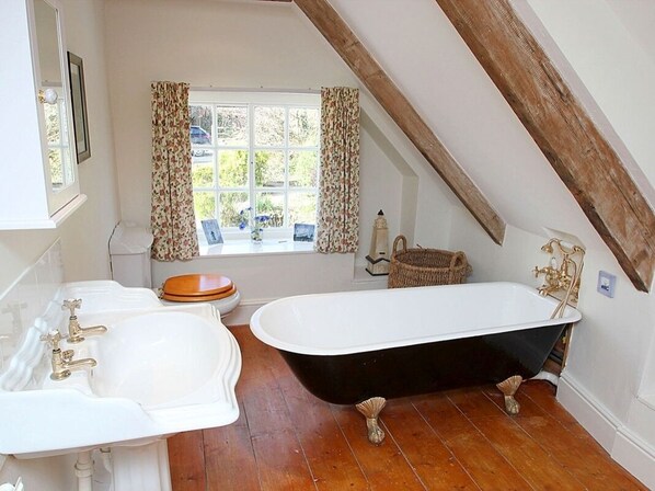 A bathroom with a freestanding bathtub, a double sink, and a window with curtains. - Rose Cottage (Helston)