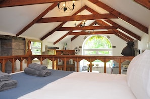 A bedroom with a high ceiling, wooden beams, a bed with a blue blanket, a stone wall, a clock, and a view of the outdoors.
