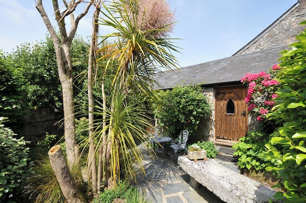 A rustic stone building with a wooden door, surrounded by lush greenery and a stone pathway.