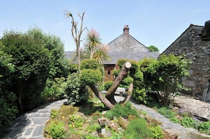 A stone pathway leading to a traditional stone building with a sloped roof, surrounded by lush greenery and a variety of plants.