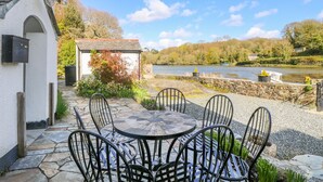A stone patio with a round table and six chairs, overlooking a river and a small white building with a bell. - Carne Mill (Helston)