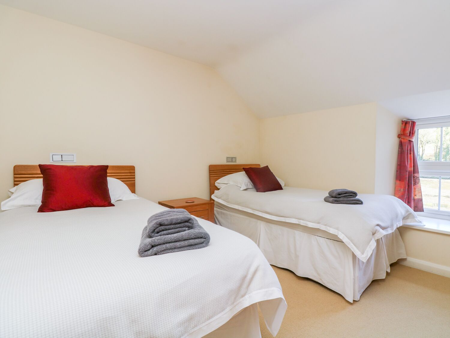 Une chambre d’hôtel avec deux lits, chacun doté d’une tête de lit en bois, de draps blancs et d’un oreiller rouge. Il y a une fenêtre avec des rideaux rouges, et une petite table avec une lampe placée entre les deux lits.
