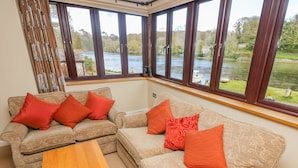 A room with two sofas and a wooden table, large windows with a view of a river and greenery. - Carne Mill (Helston)
