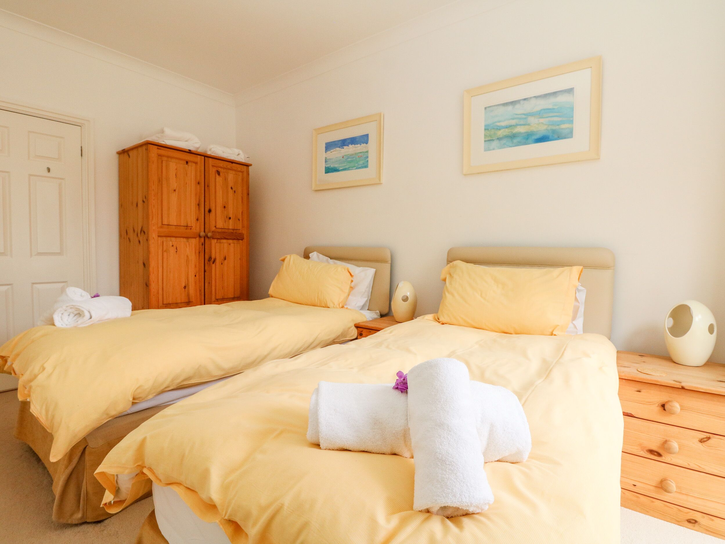 Habitación doble con dos camas, cada una con ropa de cama amarilla y toallas blancas. Se observa un armario de madera y dos cuadros en la pared.