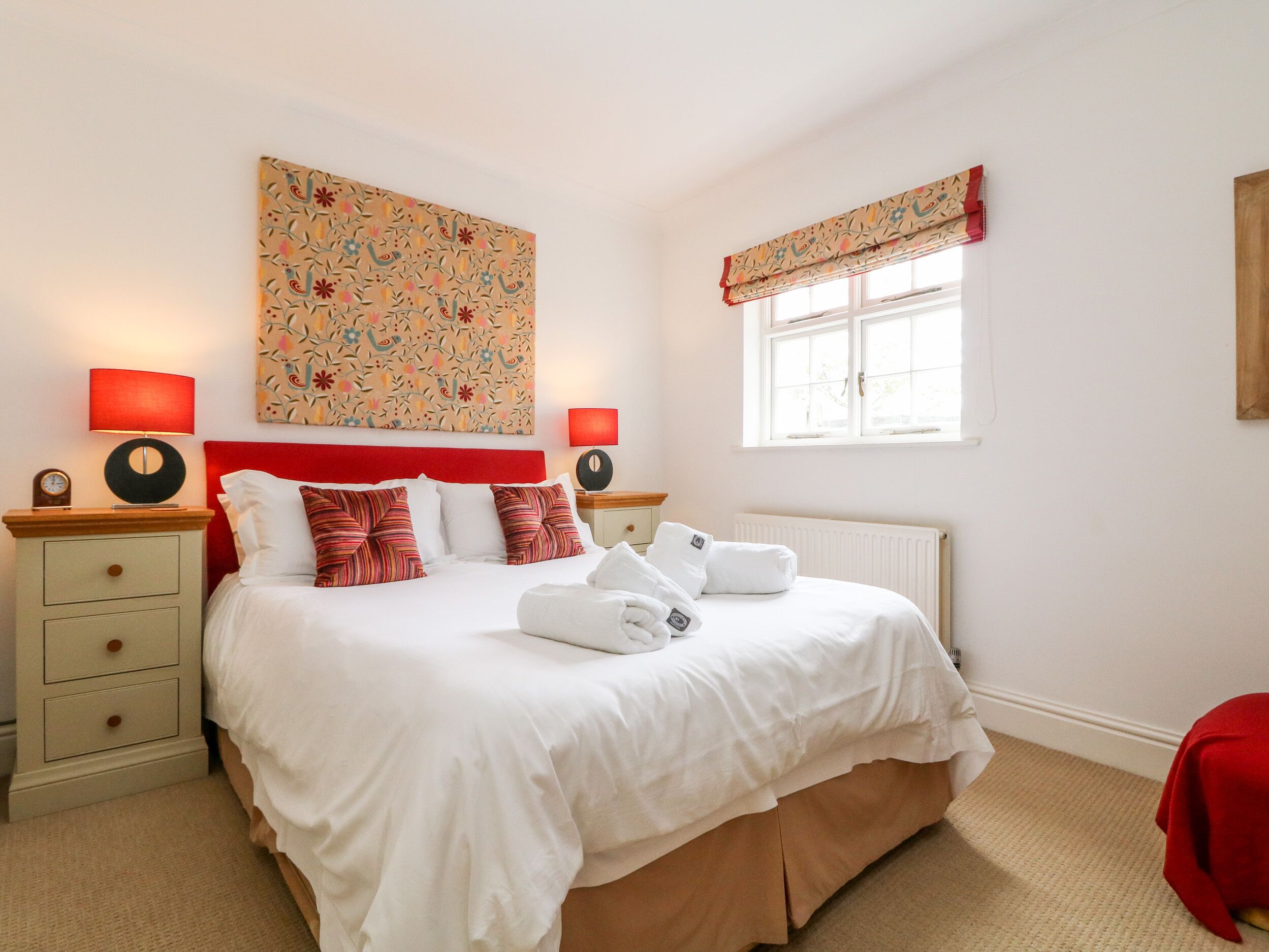 A hotel room with a bed, two bedside tables, a nightstand, a window with curtains, and a radiator.