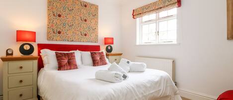 A hotel room with a bed, two bedside tables, a nightstand, a window with curtains, and a radiator.