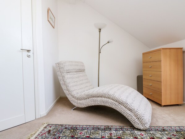 A room with a herringbone-patterned chair, a wooden dresser, a floor lamp, and a patterned rug. - Riverview (Lostwithiel)