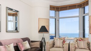 Un salon avec un canapé en cuir marron, des fauteuils beiges et une table basse en bois. Une grande fenêtre offre une vue sur la mer.