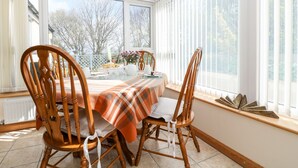 A dining area with a table set for two, wooden chairs, a plaid tablecloth, and a view of trees through large windows. - Mynheer Farm Bowji (Redruth)