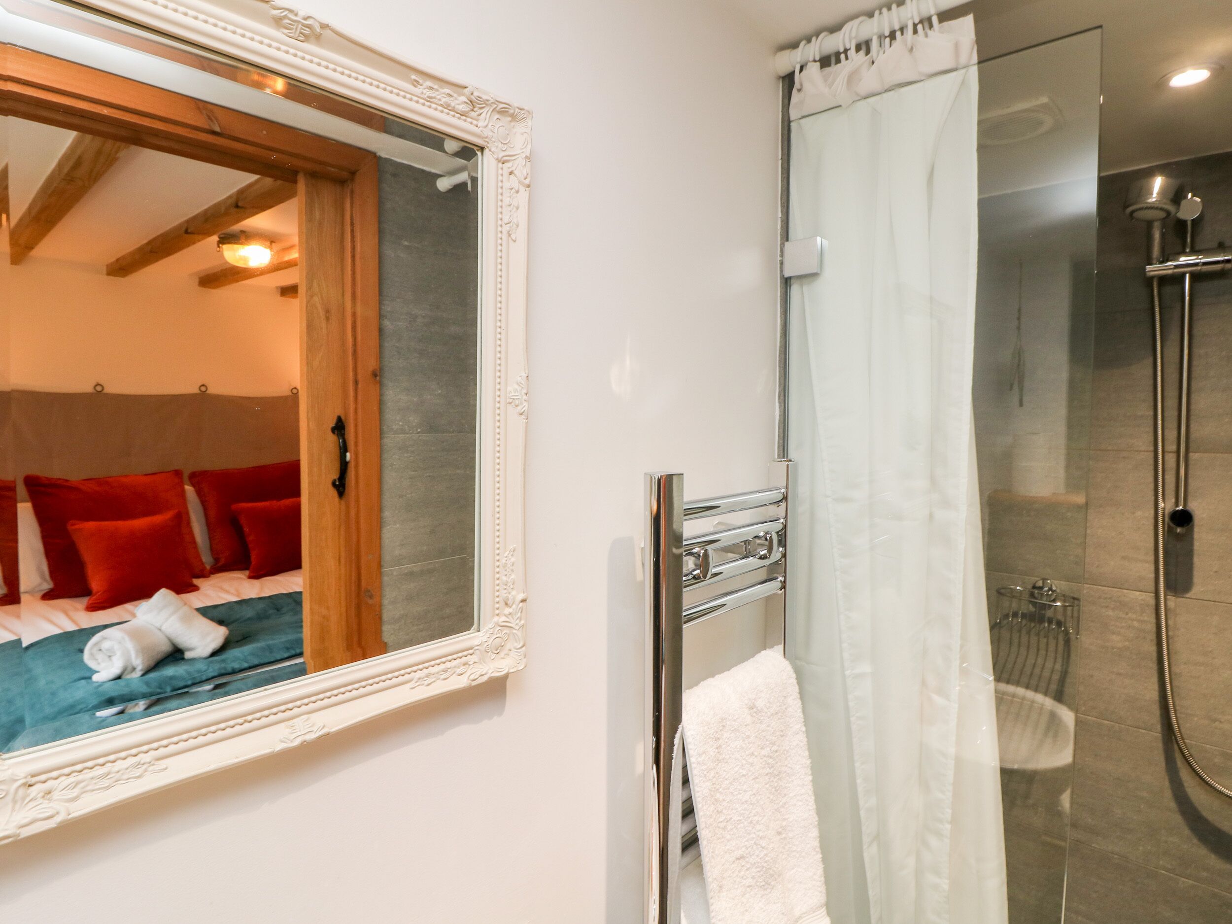 A modern bathroom with a glass shower enclosure, a white towel hanging on a rack, and a wooden door leading to a bedroom with red pillows.