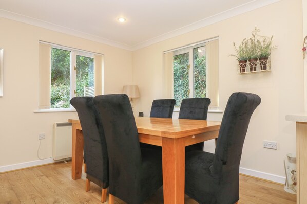 A dining area with a wooden table and four black chairs, large windows with curtains, a radiator, and a wall-mounted plant shelf. - Bramble Cottage (Falmouth)