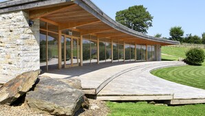 A modern building with a curved wooden deck, stone walls, and large glass windows. - Marhayes (Camelford)