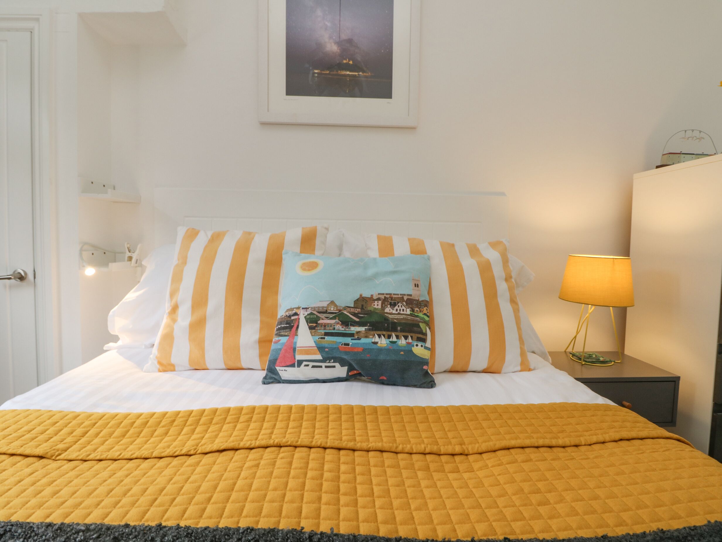 Ein ordentlich bezogenes Bett mit einer gelben Decke, gestreiften Kissen und einem Zierkissen mit Meeresmotiv. Auf dem Nachttisch neben dem Bett stehen eine Lampe und ein gerahmtes Bild.