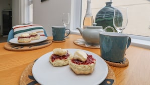 Una mesa con scones, una tetera y una botella de vino, frente a una ventana con vistas a una casa.