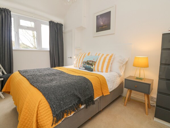 A bedroom with a bed, a bedside table, a lamp, a framed picture on the wall, and a window with curtains. - Mackerel Sky (Marazion)