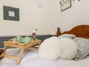 A bedroom with a wooden bed, white fluffy pillows, a bedside table with a green teapot, a vase with flowers, and a framed picture on the wall. - Little Beach House (St Austell)